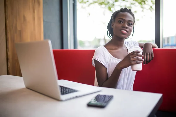 Foto de la hermosa mujer bebiendo batido sentado en la cafetería. Mujer afroamericana joven sentada en una cafetería y trabajando en un portátil . — Foto de Stock