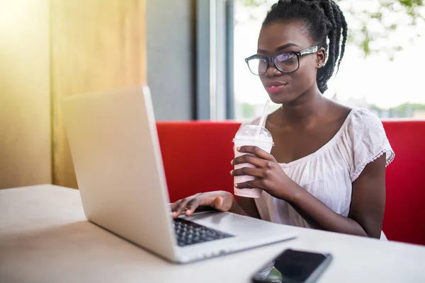 Joven afroamericana mujer beber batido sentado en una cafetería y trabajando en portátil . — Foto de Stock