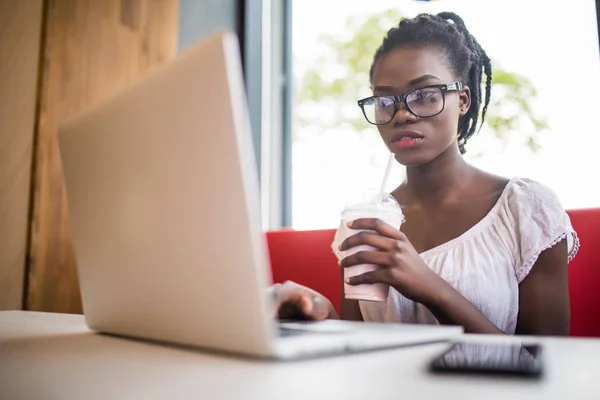 Joven afroamericana mujer beber batido sentado en una cafetería y trabajando en portátil . — Foto de Stock