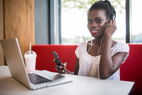 Joven chica afroamericana sentada en un restaurante con laptop. Chica bonita con auriculares mirando alegremente en el ordenador portátil en la cafetería . — Foto de Stock
