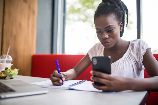 Retrato de la alegre mujer estudiante africana hacer aviso de teléfono sentado en la cafetería cerca de la universidad, lectura de resumen académico, beber café en la cafetería — Foto de Stock