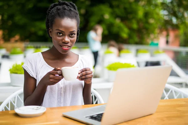 Encantadora joven empresaria afroamericana está sentada en el restaurante de la calle y tiene una sesión de video en línea remota con colegas que usan computadora portátil, cálido día soleado — Foto de Stock