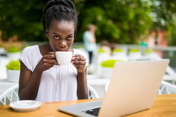 Retrato de la mujer africana usando el ordenador portátil en un café al aire libre y una taza de café en la mesa — Foto de Stock