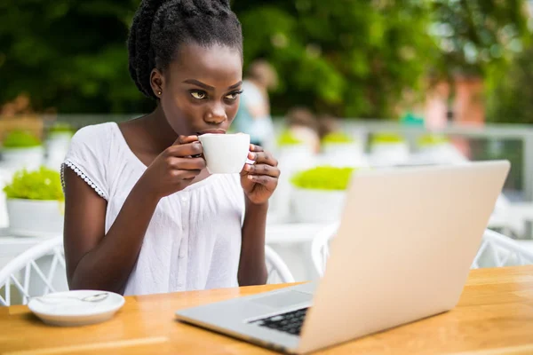 Retrato de chica afroamericana en vasos sentados en la mesa de la cafetería con portátil y sosteniendo la taza de café en las manos . — Foto de Stock