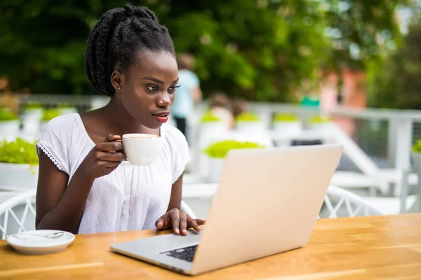 Портрет африканской женщины с ноутбуком в открытом кафе и чашкой кофе на столе — стоковое фото