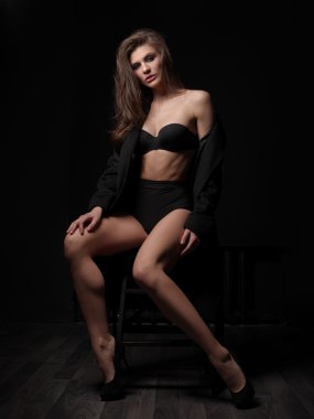 Seductive, beautiful and attractive young woman with loose brown hair, sexy gorgeous figure and long legs in the black seamless underwear and wool coat is posing on the chair in the studio clipart