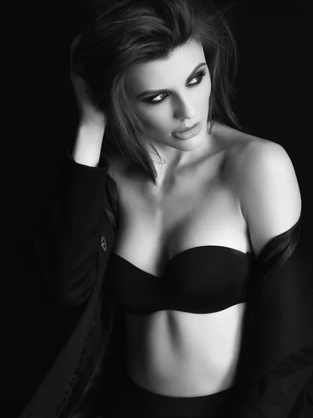 Baştan çıkarıcı, güzel ve çekici genç kadın gevşek kahverengi saçlı ve siyah dikişsiz iç çamaşırı ve yün ceket seksi rakam ile koyu arka plan, monokrom siyah ve beyaz fotoğraf poz