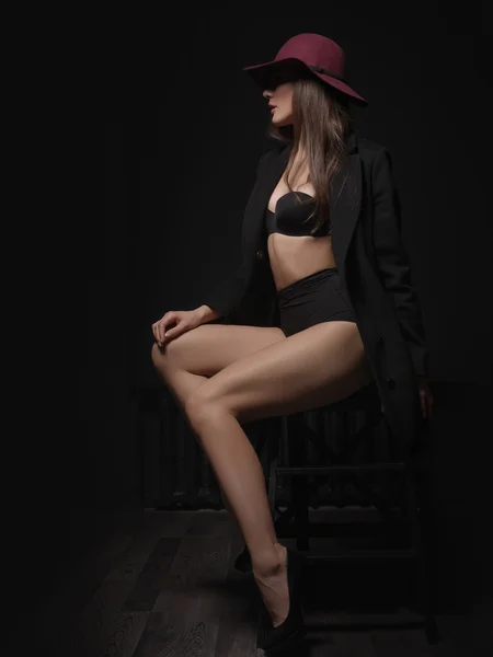 Appealing, elegant and attractive mysterious woman with loose brown hair, sexy gorgeous figure and long legs in the black seamless underwear and wool coat is posing on the chair in the studio — Stock Photo, Image