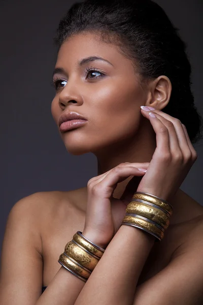 Fashionable portrait of an extraordinary beautiful naked african American female model with perfect smooth glowing mulatto skin, makeup, full lips and large golden wrist bracelets, studio photoshoot — стоковое фото