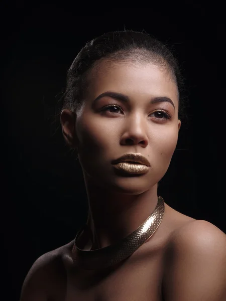 Portrait of an extraordinary beautiful naked african american model with perfect smooth glowing mulatto skin, make up, full golden lips, shaved haircut and gold jewelry on her neck, dark background