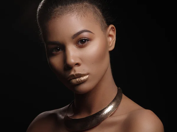 Retrato de una extraordinaria hermosa modelo afroamericana desnuda con perfecta piel mulata brillante suave, maquillaje, labios dorados completos, corte de pelo afeitado y joyas de oro en su cuello, fondo oscuro — Foto de Stock