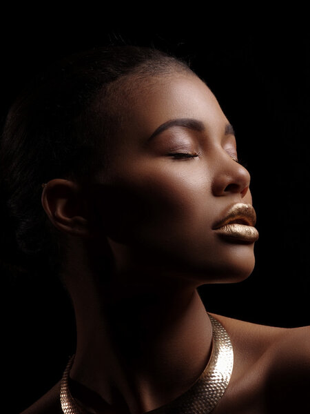 Fashion studio portrait of an extraordinary beautiful nude african american model with perfect smooth glowing mulatto skin, make up, full golden lips, shaved haircut and gold necklace, profile