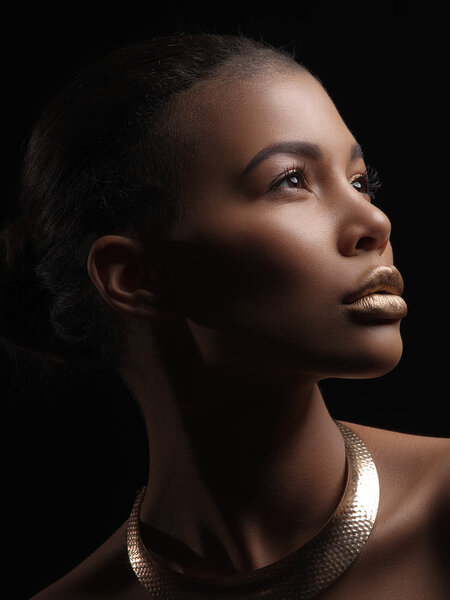 Fashion studio portrait of an extraordinary beautiful nude african american model with perfect smooth glowing mulatto skin, make up, full golden lips, shaved haircut and gold necklace, profile