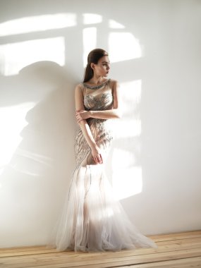 Düşünceli genç güzel kadın zarif bej Abiye ışık odasında tarafından payetler işlemeli akıllı giyinmiş ve boncuk tren ile duvara yaslanmış