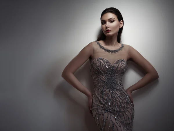 Fatale brunette with expressive dark make up and sleeked hair is posing in the studio on the gray background in the smart beige evening dress embroidered by sequins and beads — Φωτογραφία Αρχείου