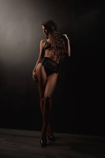 Sensual e atraente bela jovem com cabelo ondulado escuro, pernas longas e corpo magro lindo está posando na lingerie translúcida rendada preta no estúdio perto da parede, fundo escuro — Fotografia de Stock