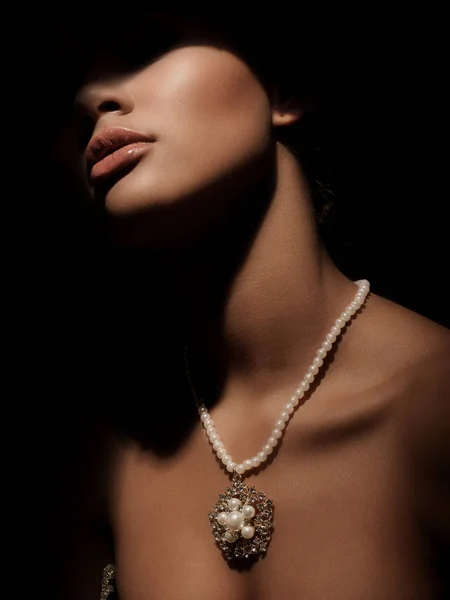 Portrait of an elegant and mystery smart lady with luxury jewelry made from precious metals on her neck (gorgeous silver necklace with pearls). Dark studio background. Shadow on the face.