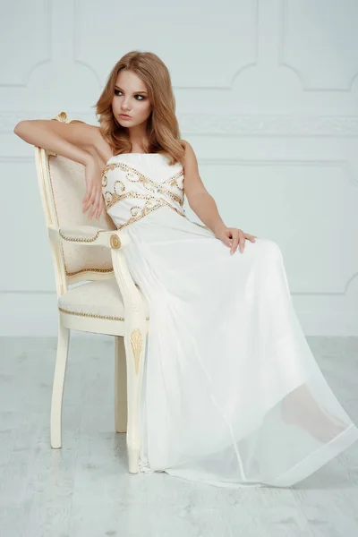 Smartly dressed charming young beautiful girl (teenager) with frail figure and blond wavy hair wearing white evening dress embroidered with sequins is posing in the light interior studio room