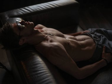 Sıcak, şık, kendine güvenen ve kendini beğenmiş maço adam atletik vücut (çıplak vücut'la seksi karın) giyen güneş gözlüğü ile deri kanepede kapalı poz  