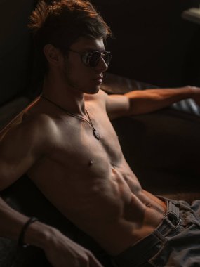 Sıcak, güzel, kendine güvenen ve yakışıklı adam atletik vücut (çıplak vücut'la seksi karın) giyen güneş gözlüğü ile deri kanepede kapalı poz  