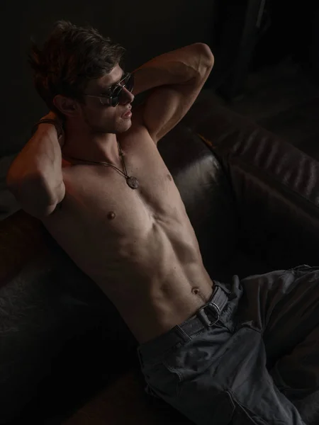 Hot, beautiful, self-confident and handsome guy with athletic body (naked torso with sexy abdominal) wearing sunglasses is posing indoors on the leather sofa