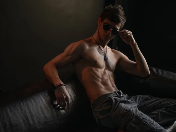 Hombre macho caliente, elegante, seguro de sí mismo y narcisista con cuerpo atlético (torso desnudo con abdomen sexy) usando gafas de sol está posando en el interior del sofá de cuero —  Fotos de Stock