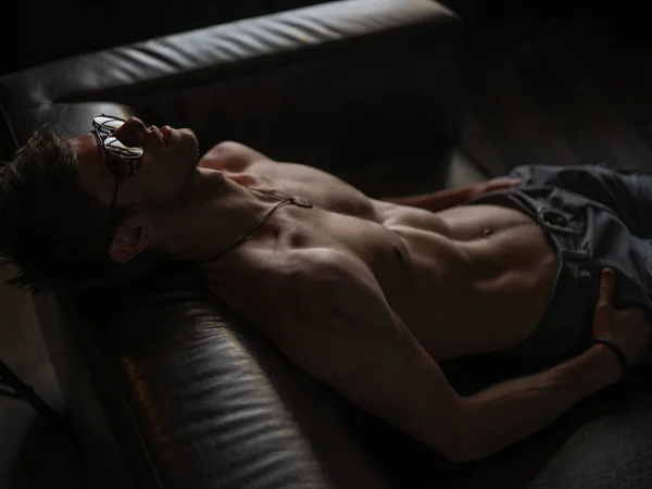 Sıcak, şık, kendine güvenen ve kendini beğenmiş maço adam atletik vücut (çıplak vücut'la seksi karın) giyen güneş gözlüğü ile deri kanepede kapalı poz  