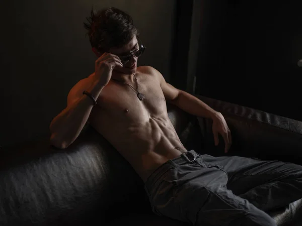 Caliente, hermoso, seguro de sí mismo y narcisista chico con cuerpo atlético (torso desnudo con sexy abdominal) usando gafas de sol está posando en el interior en el sofá de cuero — Foto de Stock
