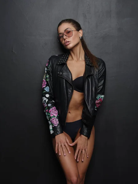 Mujer joven seductora, hermosa y atractiva con sexy figura caliente delgada está posando en la ropa interior sin costuras negro, chaqueta de cuero impresa con estilo y chaqueta de gafas de sol cerca del fondo oscuro en el estudio — Foto de Stock