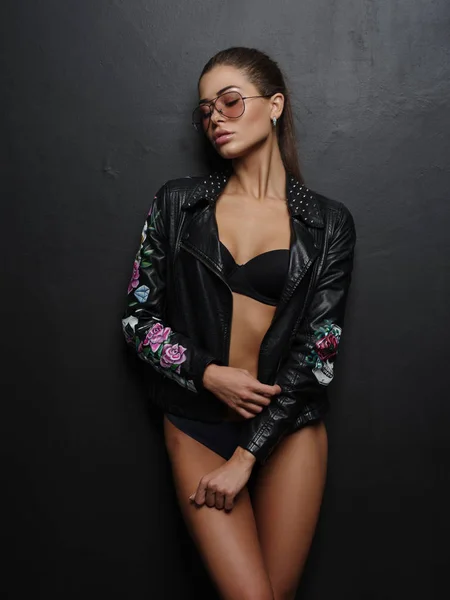 Verführerische, schöne, attraktive Mädchen mit sexy wunderschönen gebräunten Körper posiert in der schwarzen nahtlosen Dessous und stilvolle Biker-Lederjacke und Sonnenbrille in der Nähe der dunklen Wand im Studio — Stockfoto