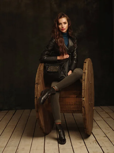 Beautiful, self-contained, confident and successful young woman is sitting and posing dressed in a stylish black leather jacket (studio shoot of catalogue of fashionable clothes, model demonstrates the collection)