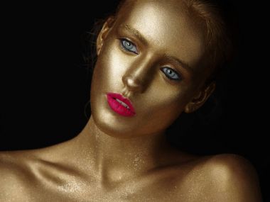 Fashionable portrait of a beautiful young female model with creative body art glowing make up. Face is colored with gold paint. Golden shine of skin. Studio black background. clipart