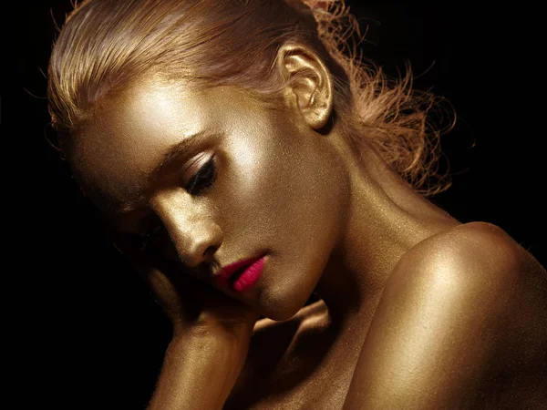 Glamorous portrait of a beautiful young woman with creative body art glowing make up. Face is painted with gold color. Golden shine of naked skin. Studio black background. — Stock Photo, Image