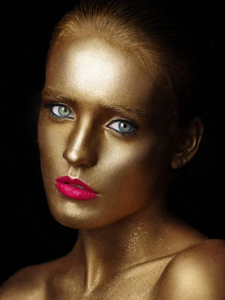 Fashionable portrait of a beautiful young female model with creative body art glowing make up. Face is colored with gold paint. Golden shine of skin. Studio black background. — Stock Photo, Image