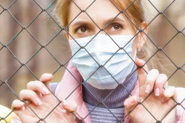 The quarantined woman in the medical mask looks through the metal mesh of the fence