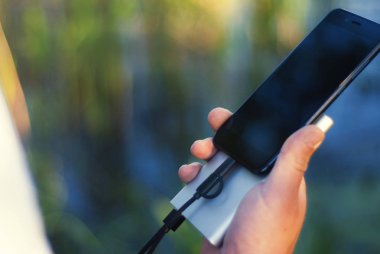 Adamın elinde akıllı telefonu olan taşınabilir bir şarj aleti var. Doğanın arka planında yeşil ve gölü olan bir adam.