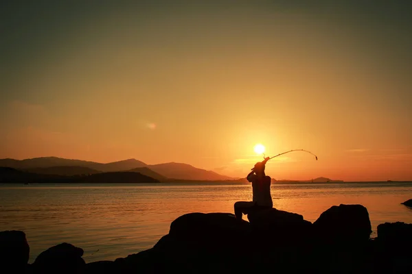 Jovem sentado em pedra e pesca no fundo do pôr-do-sol mar — Fotografia de Stock