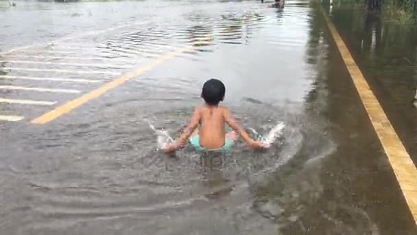 Щасливий азіатський хлопчик грає воду на дорозі, затоплена дорога, після сильного дощу ввечері — стокове відео