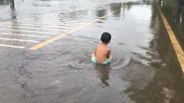 Счастливый азиатский мальчик играть в воду на дороге, затопленной дороге, после сильного дождя вечером — стоковое видео