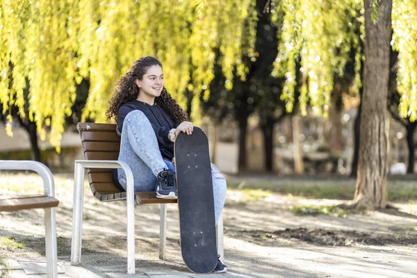 Adolescente bonito sentado em um banco com seu skate bordo — Fotografia de Stock