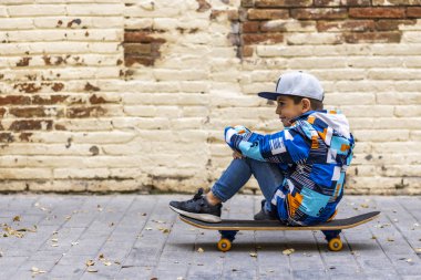 Kaykayının üstünde oturan sevimli küçük çocuk, arkasında tuğla bir duvarla.