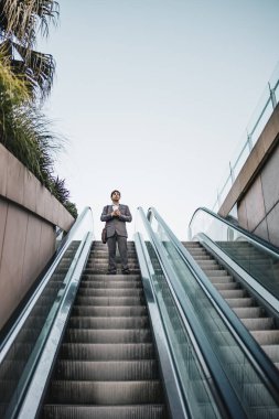 Takım elbiseli bir işadamı kahve içiyor ve yürüyen merdivenden çıkıyor.