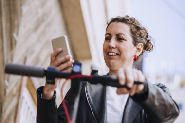 Orta yaşlı bir kadın elektrikli scooterına biniyor ve cep telefonuna bakıyor..