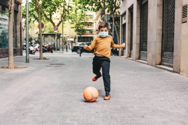 Küçük çocuk maskeyle futbol oynuyor.