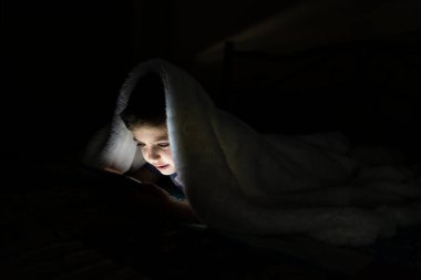 Küçük çocuk battaniyeyle kaplı bir tablet kullanıyor.