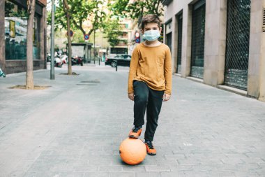 Küçük çocuk maskeyle futbol oynuyor.