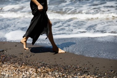 Deniz kenarında dalgalı siyah bir elbiseyle kumda yürüyen çıplak ayaklı bir kız.