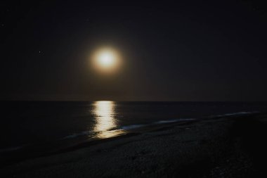 Deniz kıyısı. Ay parlıyor ve yıldızlar görünüyor. Kıyı şeridini de görebilirsiniz. Gece.