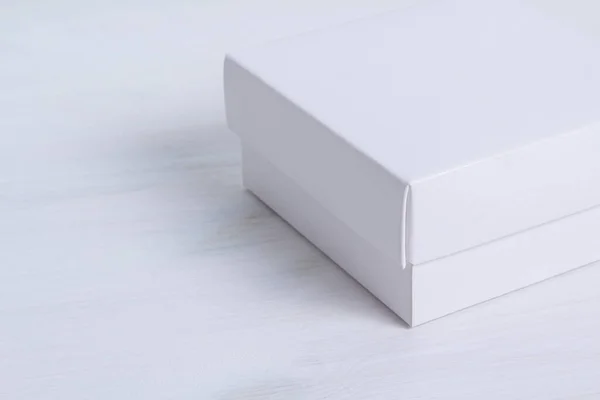 木製の背景に蓋付きの白いギフトボックスを閉じます。クロップ画像、サイドビュー、正方形のボックス。エレガントなエコフレンドリーなモックアップ。白い木製のテーブルの上にカバー付きの隔離された箱。左側の空のスペース. — ストック写真