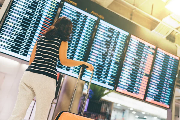 Asian woman traveler looking at flight information screen in an airport, holding suitcase, travel or time concept, warm light effect — Stockfoto